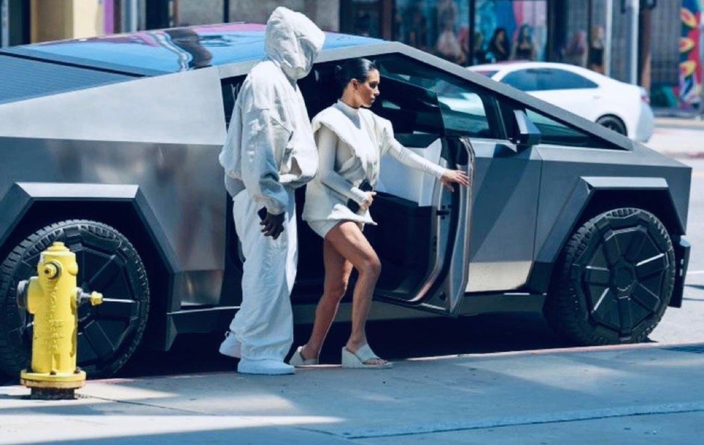 A person in a white outfit steps out of a futuristic gray vehicle, while another person in a matching white outfit stands nearby. A yellow fire hydrant is visible in the foreground.
